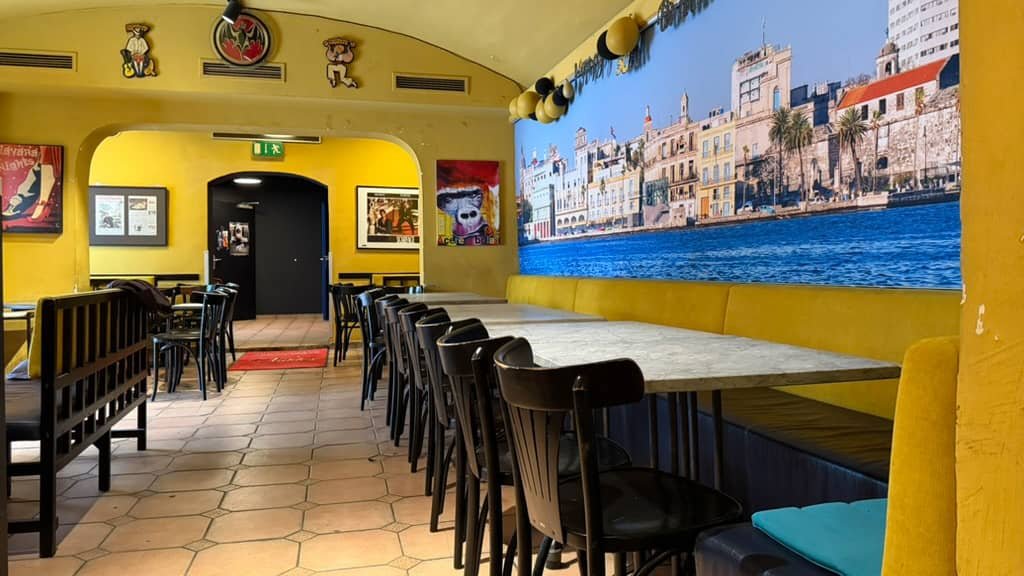 Gelber Gastraum mit langen Tischen und einem großen Wandbild einer Küstenstadt.