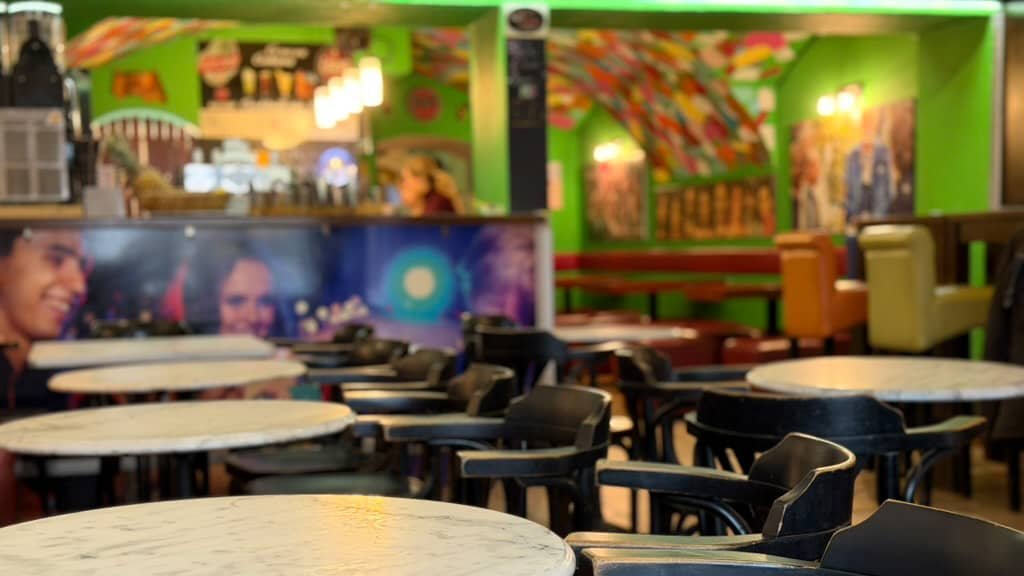 Zahlreiche runde Marmortische mit dunklen Holzstühlen in einem Café. Blick über die runden Tische auf eine farbenfrohe Bar mit grünen Akzenten.