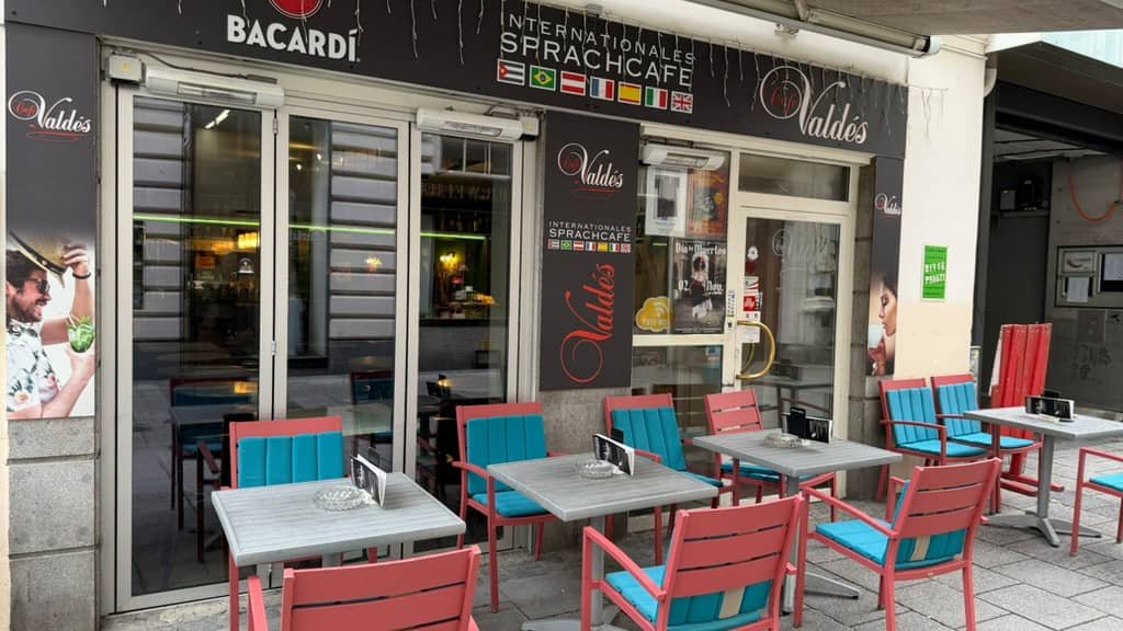 Außenansicht Café: Eingangsbereich des „Café Valdés“ mit bunten Stühlen und Tischen auf dem Gehweg.