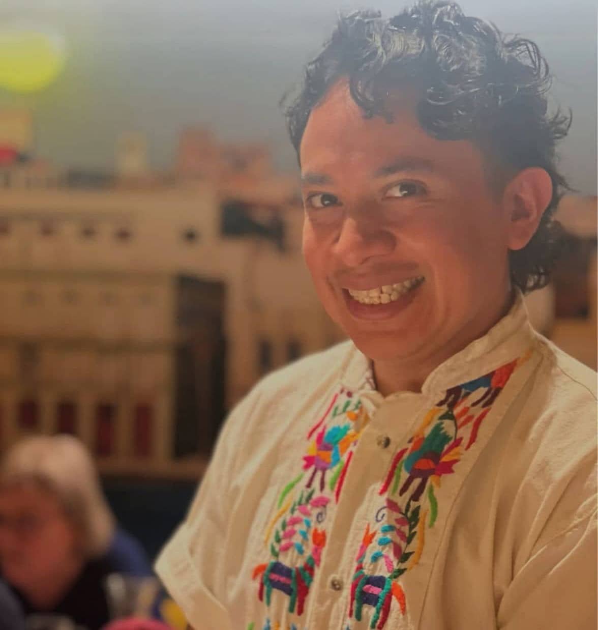 „Lächelnde Person mit lockigem Haar in einem hellen, bestickten Hemd vor unscharfem Innenraum.“