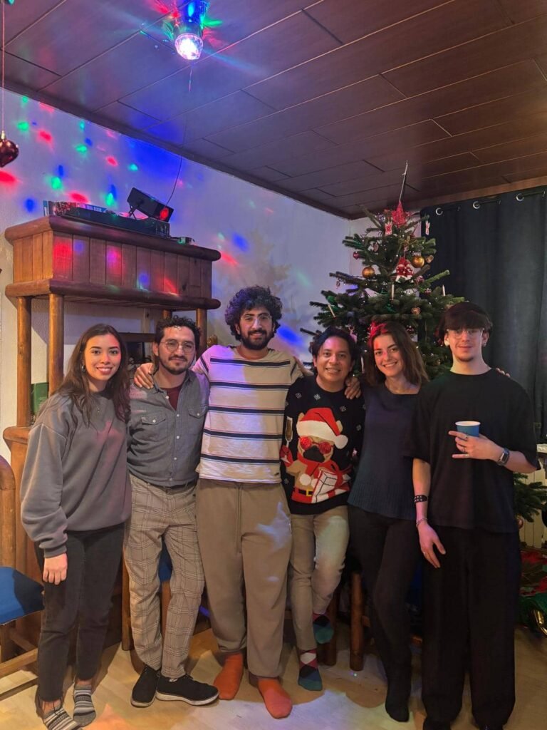 Sechs lächelnde junge Erwachsene posieren Arm in Arm in einem festlich dekorierten Raum. Im Hintergrund sind ein geschmückter Weihnachtsbaum und bunte Party-Lichter an der Wand zu sehen.