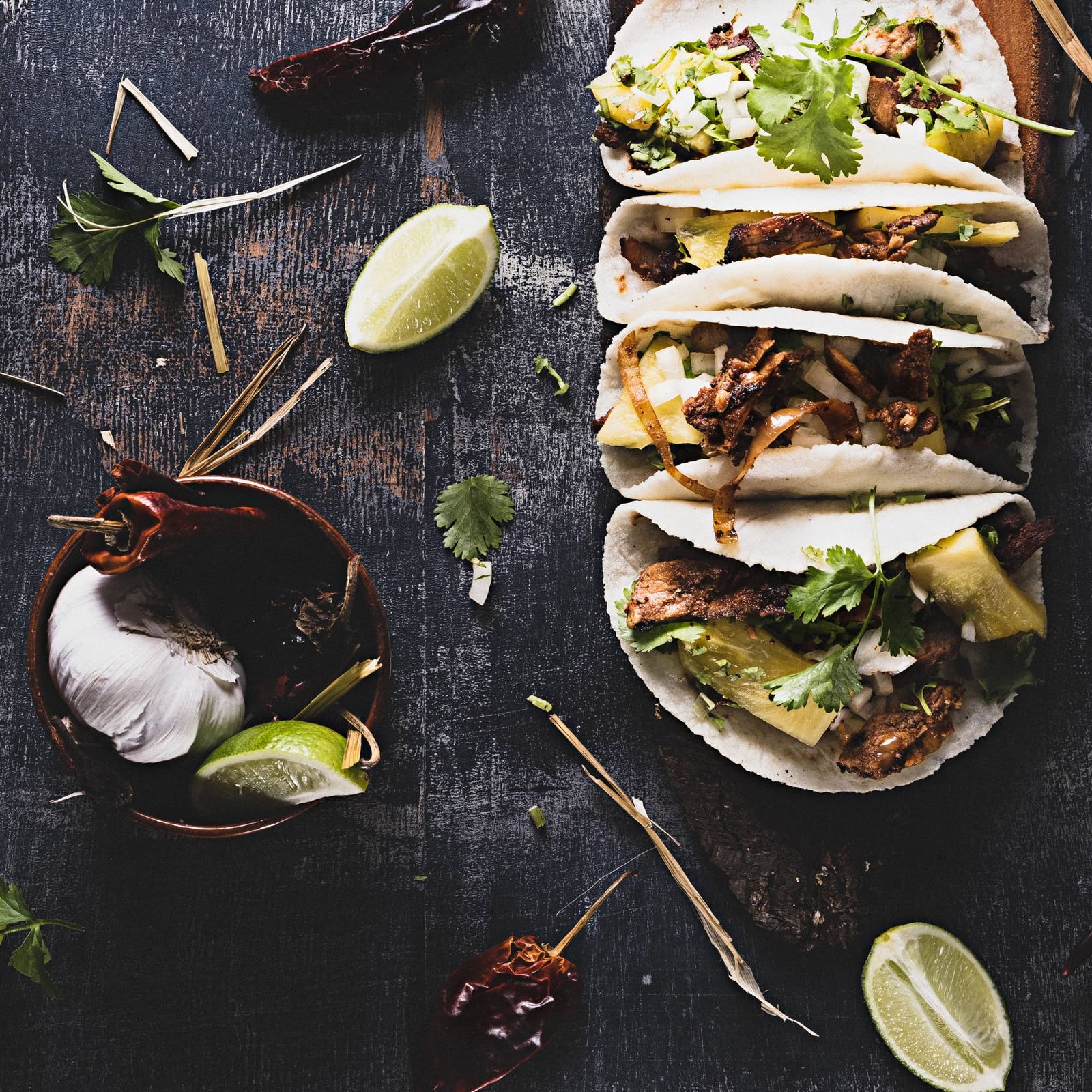 Drei gefüllte Tacos mit gegrilltem Fleisch, Ananas, Zwiebeln und frischem Koriander liegen auf einer dunklen Holzoberfläche; rundherum sind Limettenspalten, Knoblauch, getrocknete Chilischoten und Kräuter dekorativ verteilt.