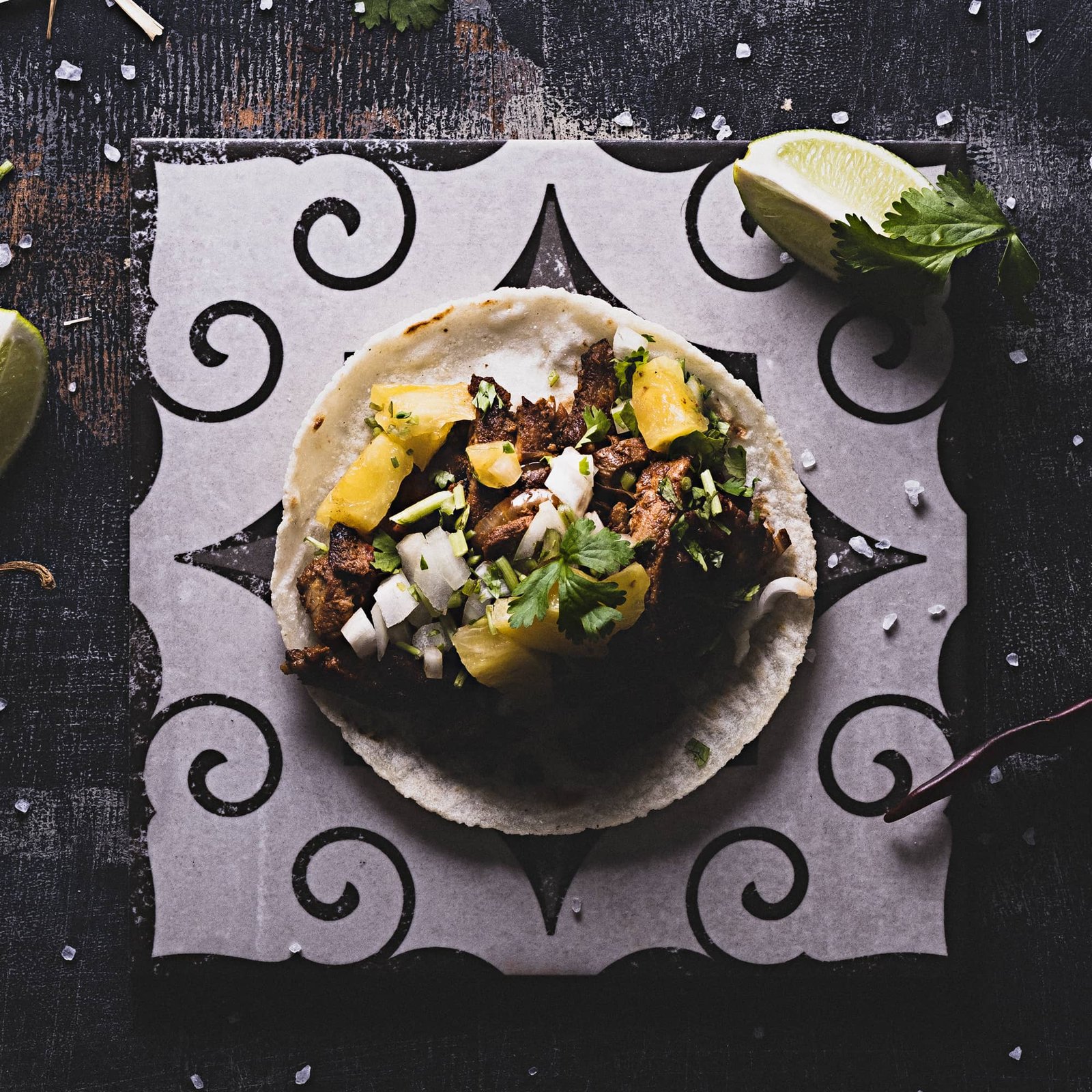 Ein einzelner Taco mit gegrilltem Fleisch, Ananasstücken, gewürfelten Zwiebeln und frischem Koriander liegt auf dekorativ gemustertem Papier; daneben sind eine Limettenspalte, grobes Salz und Chilischoten auf einer dunklen Oberfläche zu sehen.