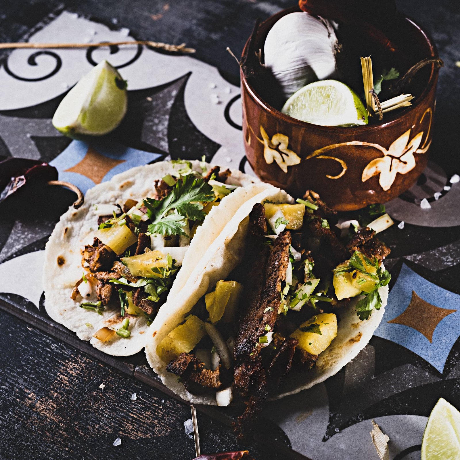 Zwei gefüllte Tacos mit gegrilltem Fleisch, Ananas, Zwiebeln und frischem Koriander liegen auf gemustertem Papier; im Hintergrund stehen eine rustikale Schale mit Knoblauch und Limette sowie grobes Salz und Chilischoten auf einer dunklen Holzoberfläche.